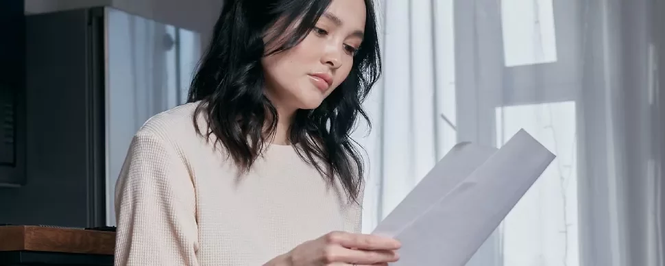 Person seated, holding and reading papers by a window.