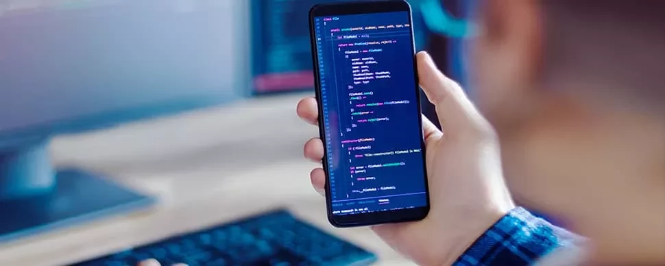 Person holding a phone displaying code, with a blurred computer screen in the background.