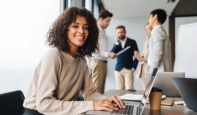 Person smiling at desk with laptop, group discussing in background.