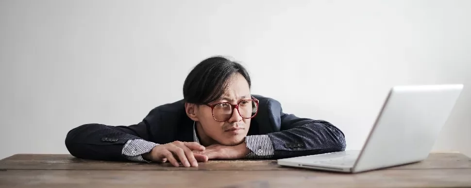 Person in a suit resting head on hands, staring at a laptop on a wooden table.