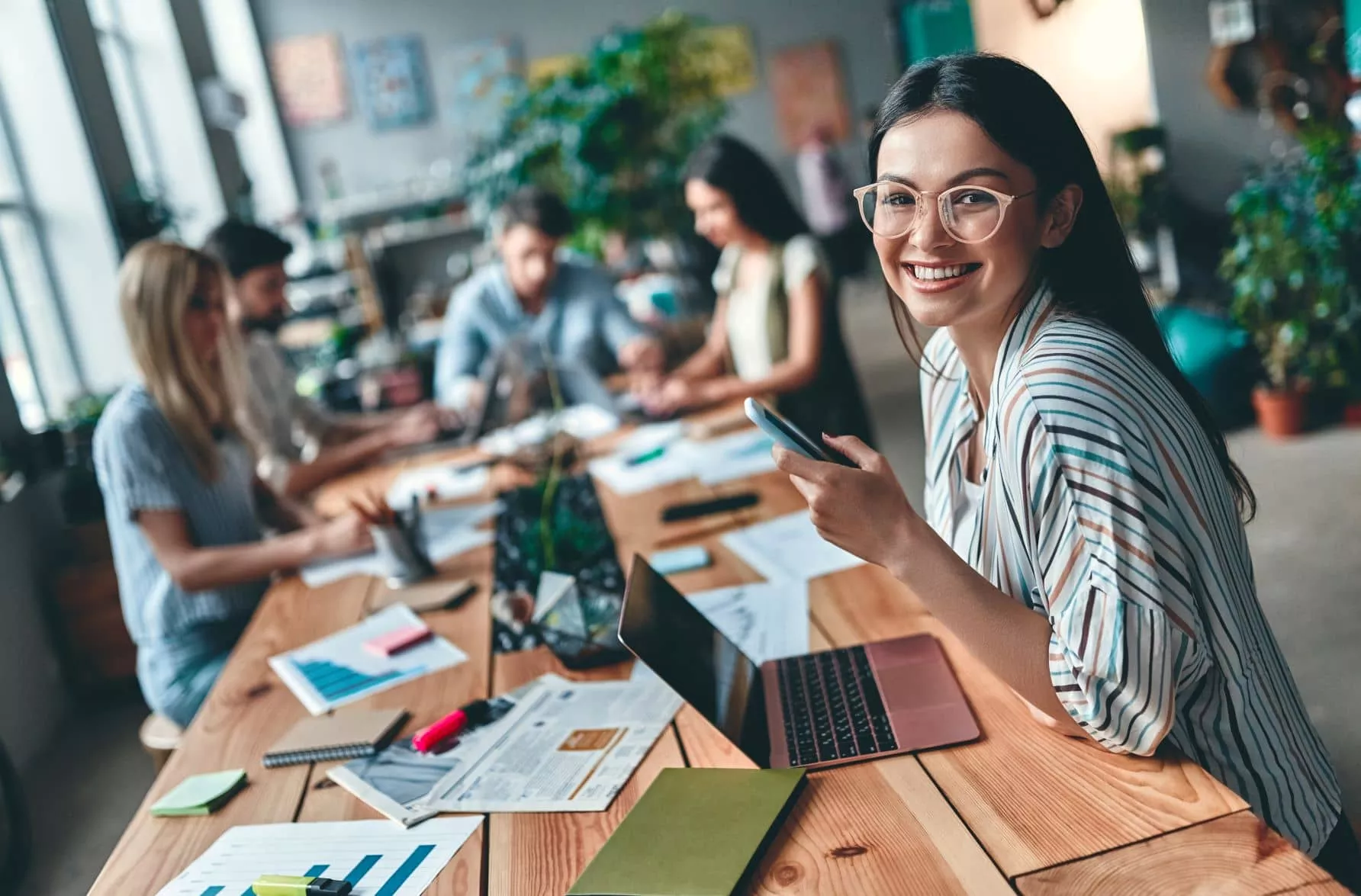 Person smiling at camera, holding phone in a meeting room with others working at a table.
