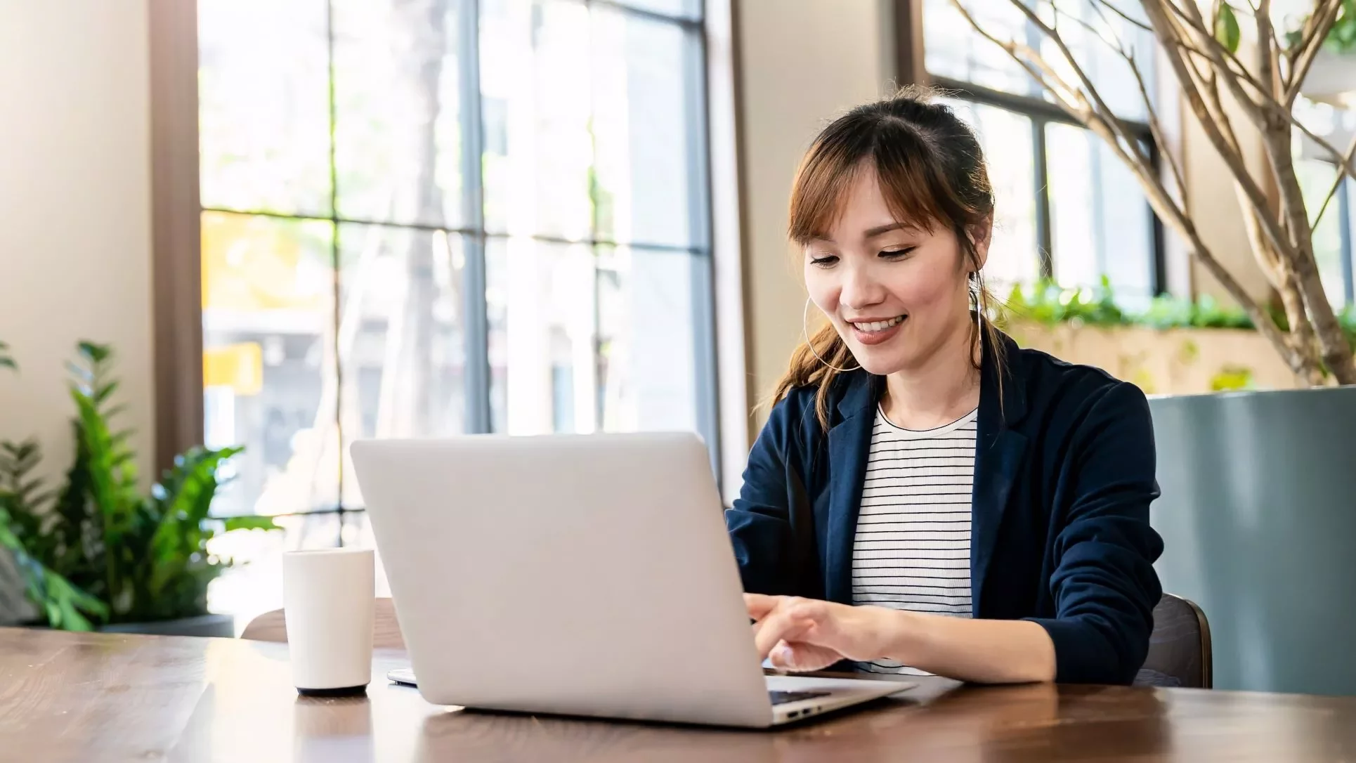Person smiling while working on a laptop at a wooden table with plants and windows in the background.