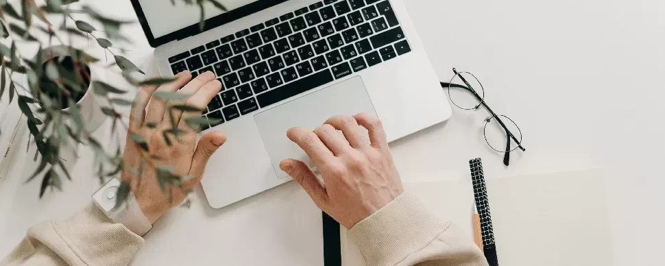Person working on a laptop at a desk with notebooks and glasses.