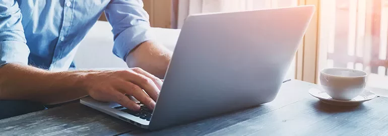 Person typing on a laptop at a wooden table with a cup of coffee beside them.