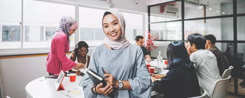 Person smiling in an office with colleagues gathered around a table.