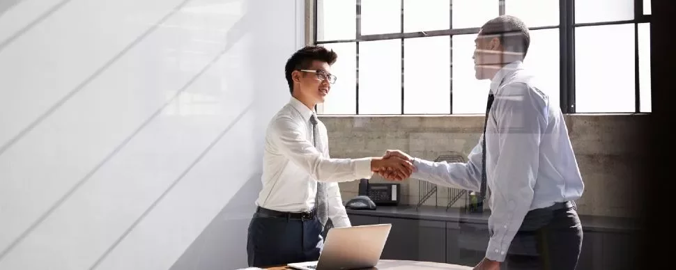 Two people in an office shaking hands by a desk with a laptop.