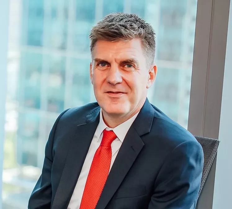 Person in a suit with red tie, sitting by a window in an office.