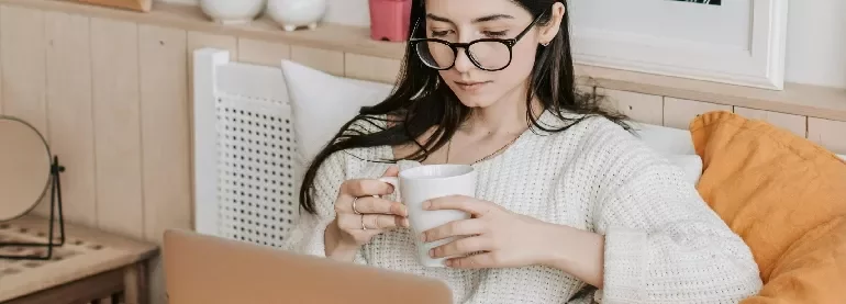 Person sitting on a cozy couch, holding a coffee mug while using a laptop.