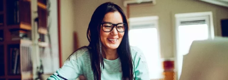 Woman in glasses smiling at a computer screen in a cozy room.