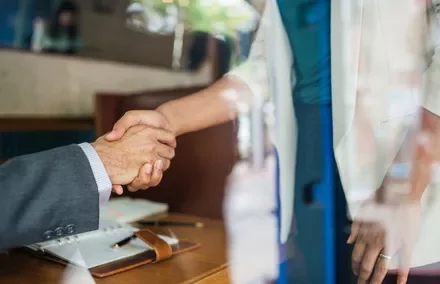 Two people shaking hands over a wooden table, seen through a window.