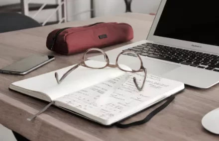 Eyeglasses resting on an open notebook with a laptop and pencil case on a wooden desk.
