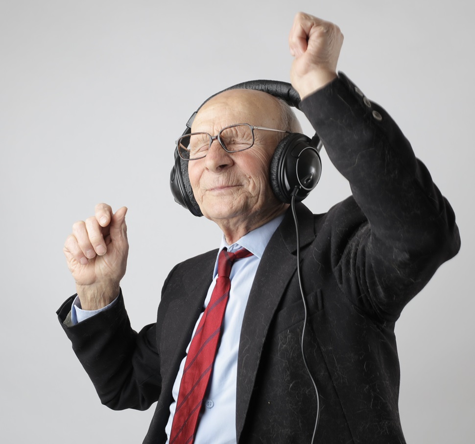 An elderly man in a work suit with red tie dancing with eyes closed, while wearing headphones.